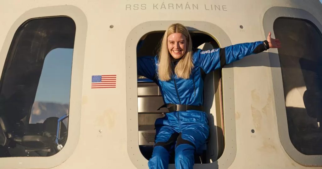 Michaella Benthaus con traje de vuelo azul sale de la escotilla de la cápsula "RSS Kármán Line" con los brazos abiertos y una gran sonrisa de entusiasmo.
