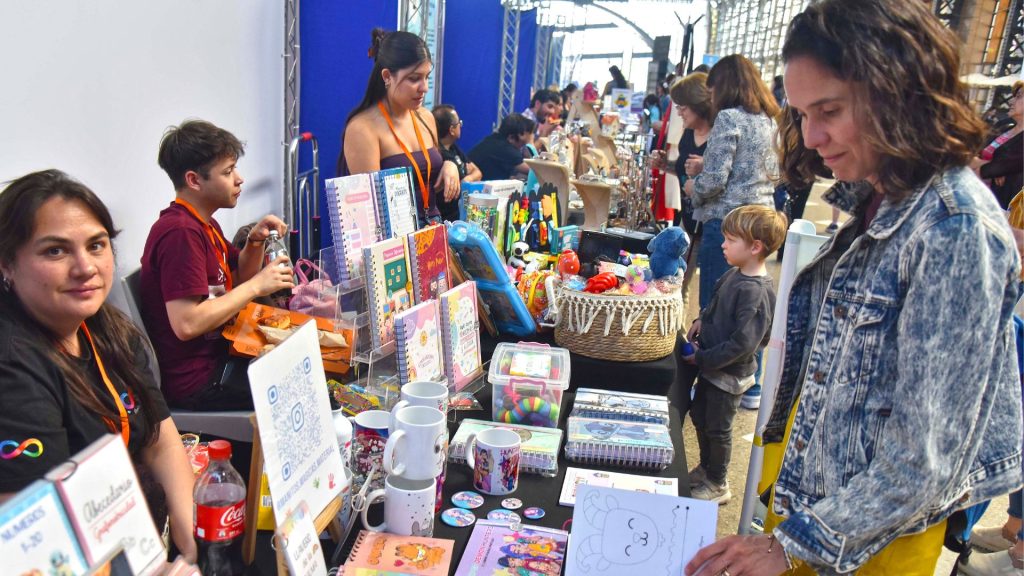 Fotografía de un animado puesto en una feria de emprendedores; vendedores atienden detrás de una mesa llena de cuadernos, tazas y regalos, mientras una mujer y un niño observan los productos.