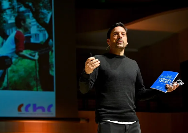 Hombre dando una conferencia en un escenario, usando un micrófono de diadema y sosteniendo el libro "Ciudades Azules" en su mano izquierda.