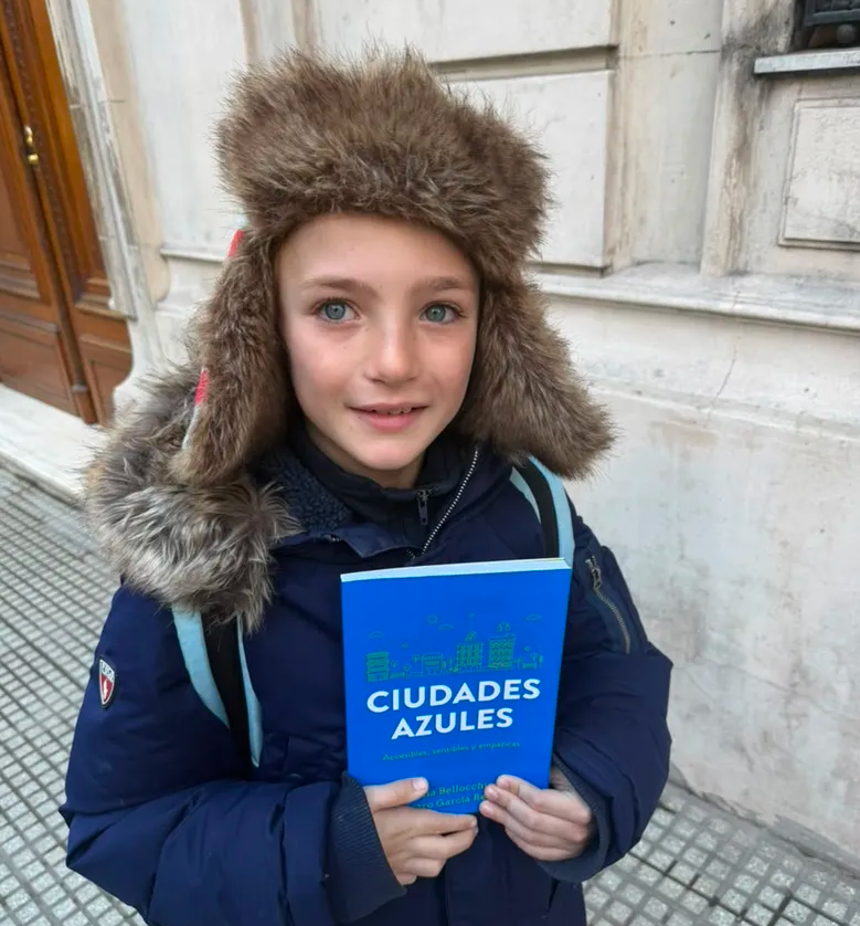 Niño sonriente con ropa de invierno y gorro de piel sosteniendo un libro azul titulado "Ciudades Azules" en la calle.